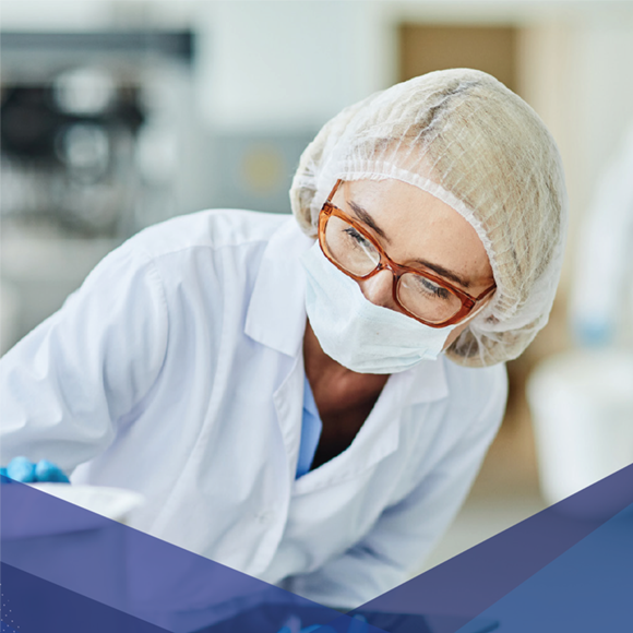 Un technicien de laboratoire portant un masque, une charlotte et des lunettes, travaillant dans un environnement clinique ou stérile.